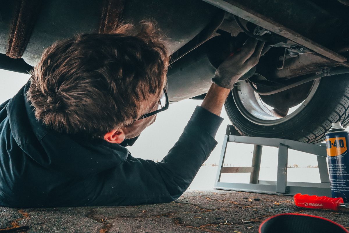Mobile-style mechanic lying under a raised vehicle doing hands-on underbody repair work outdoors