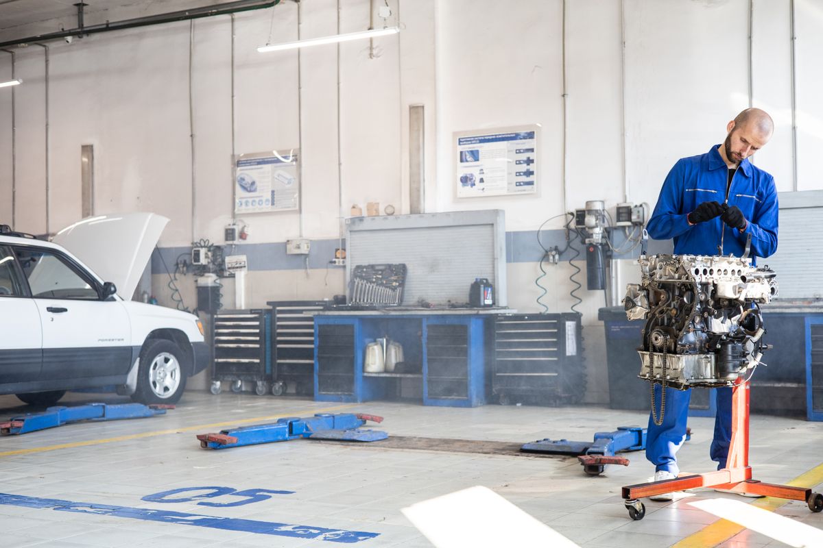 Mechanic in blue coveralls working on a vehicle engine on a stand inside an ordinary trade workshop, with a white SUV on a hoist